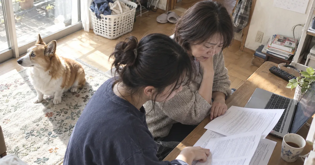 机で書類を読む母親と成人した娘と、ラグの上に座るコーギー