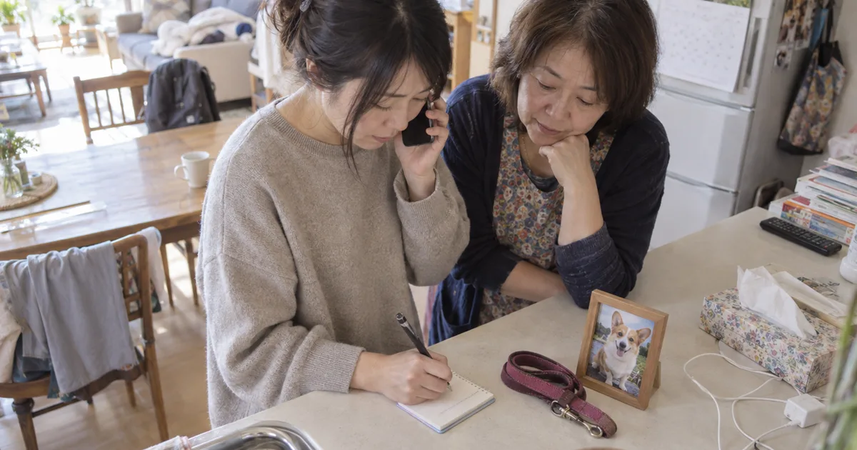 キッチンカウンターで電話をしながらメモを取る母親と成人した娘と、コーギーの写真とリード